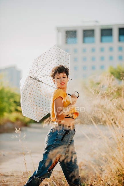 Молодая женщина в стильном наряде смотрит в сторону, идя под капли воды с зонтиком — стоковое фото