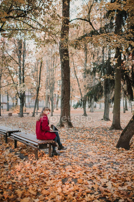 Молода пані в червоному пальто за допомогою пристрою і сидячи на сидінні в осінньому лісі. — Stock Photo