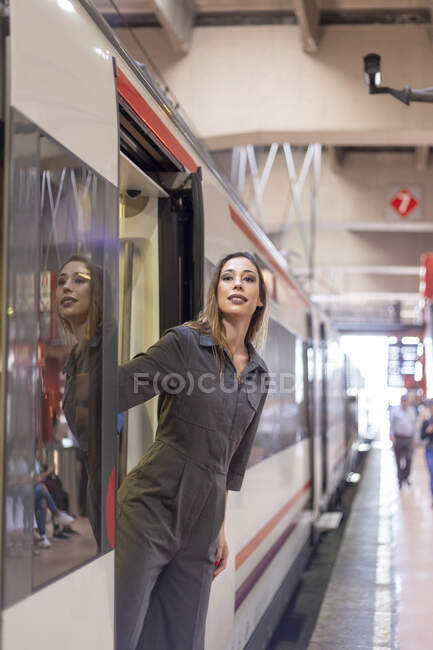 Hermosa hembra en traje elegante sonriendo y mirando hacia otro lado mientras se inclina hacia fuera tren moderno en la estación de tren - foto de stock