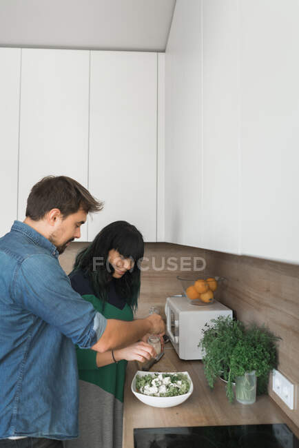 Vista lateral de cara jovem bonito e linda mulher cozinhar salada saudável, enquanto de pé na cozinha elegante juntos — Fotografia de Stock