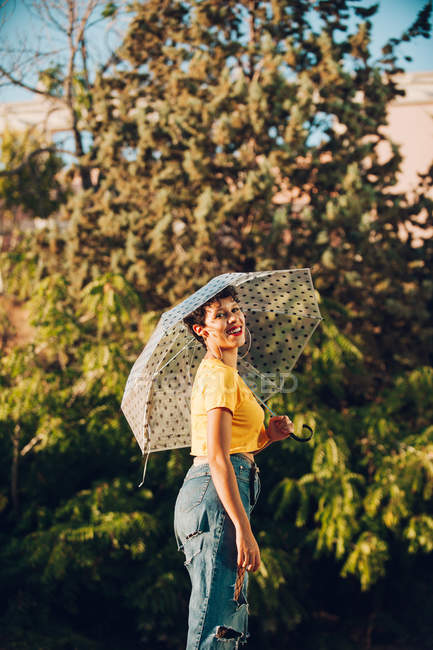 Молода жінка в стильному повсякденному вбранні дивиться на камеру, тримаючи парасольку і стоячи на вулиці біля кущів — стокове фото