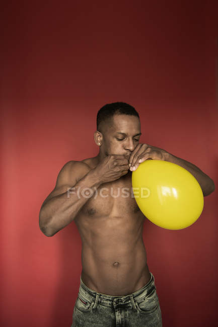 Musculoso hombre negro sin camisa en jeans soplando globo amarillo brillante sobre fondo rojo - foto de stock