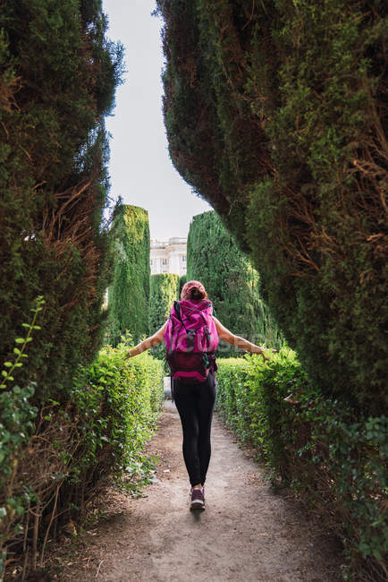 Visão traseira de esportista com mochila rosa andando no parque entre arbustos verdes exuberantes à luz do dia — Fotografia de Stock