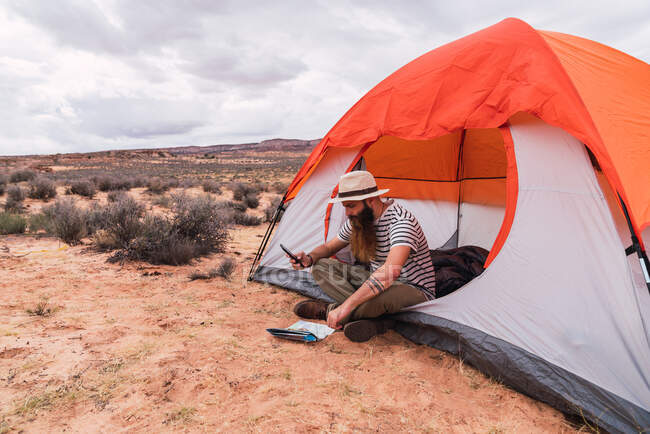 Веселый парень в повседневной одежде со смартфоном и картой, сидя возле палатки и глядя вдаль на красивую природу — стоковое фото