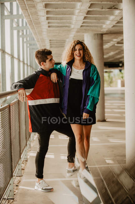 Cheerful young woman smiling and leaning on shoulder of handsome guy while standing under roof in modern pathway — Stock Photo