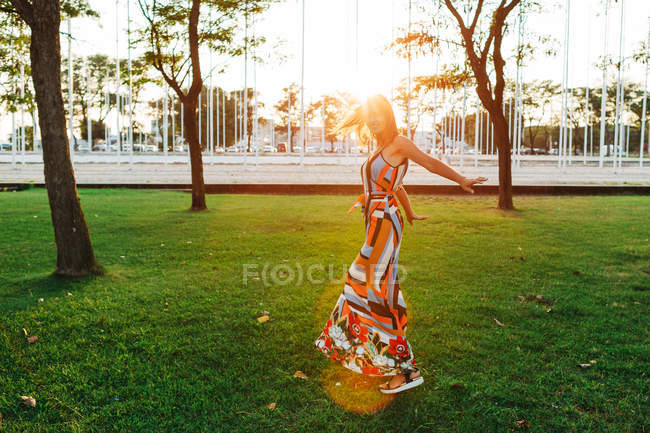 Morena elegante alegre em vestido girando em torno do prado verde no parque urbano — Fotografia de Stock