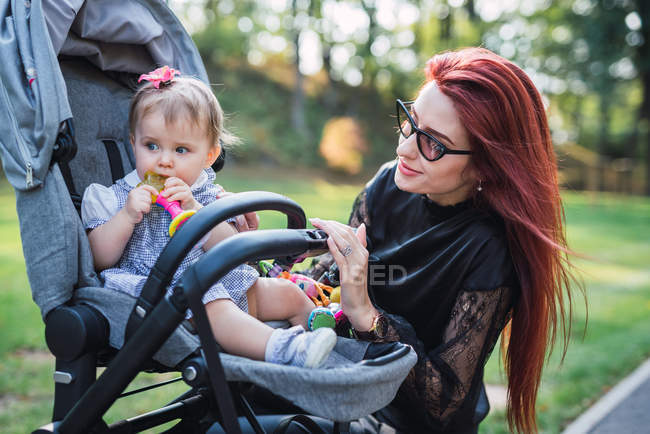 Junge Mutter sieht Mädchen im Kinderwagen im sonnigen Park an — Stockfoto