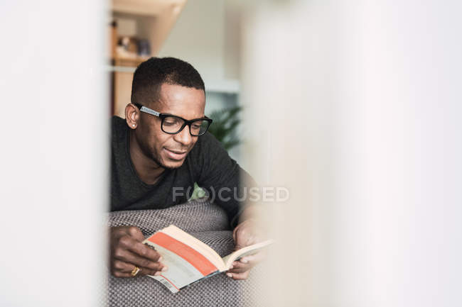 Сосредоточенный афроамериканец в очках читает книгу, сидя дома на диване — стоковое фото