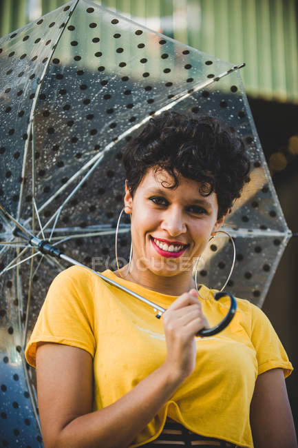 Retrato de mujer joven con paraguas transparente sonriendo y mirando a la cámara mientras está de pie en la calle - foto de stock