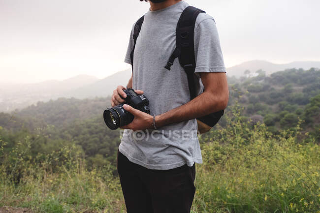 Unrecognizable guy in casual outfit holding professional photo camera while standing in nature — Stock Photo
