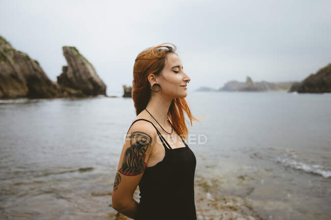 Vista laterale di bella giovane donna tenere gli occhi chiusi e godersi il tempo mentre in piedi vicino al mare su Playa de Arnia in Spagna — Foto stock