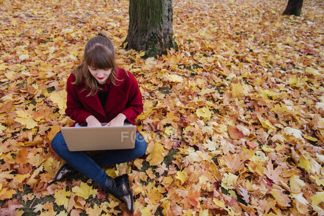 Side view of lady in red coat using?device and sitting on soil between falling yellow maple leaves — Stock Photo