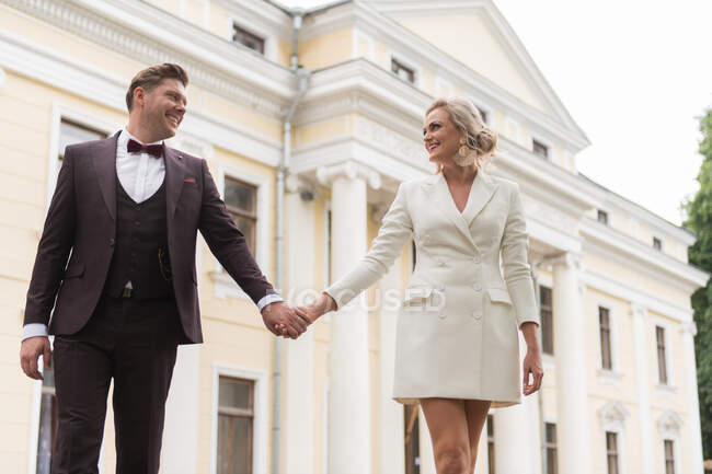 Знизу красива жінка і чоловік в елегантних весільних костюмах і посміхається один одному на тлі старої будівлі — стокове фото