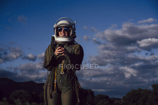 Smiling female astronaut in sunglasses holding mobile phone against evening sky — Stock Photo