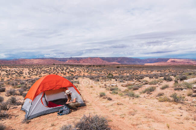 Vista lateral de cara bonito em roupa casual mochila de embalagem enquanto sentado no chão perto da barraca na área de acampamento no deserto — Fotografia de Stock