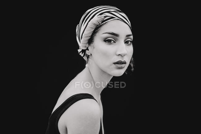Vista laterale di bella giovane donna in tessuto testa a righe con trucco luminoso guardando la fotocamera isolata su sfondo nero — Foto stock