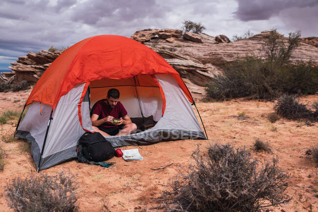 Crop man con ciotola di insalata fresca seduto alla tenda moderna sulla zona campeggio nel deserto — Foto stock