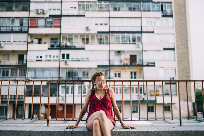 Menina ruiva elegante com tranças e óculos de sol sentado perto de trilhos contra edifício residencial — Fotografia de Stock