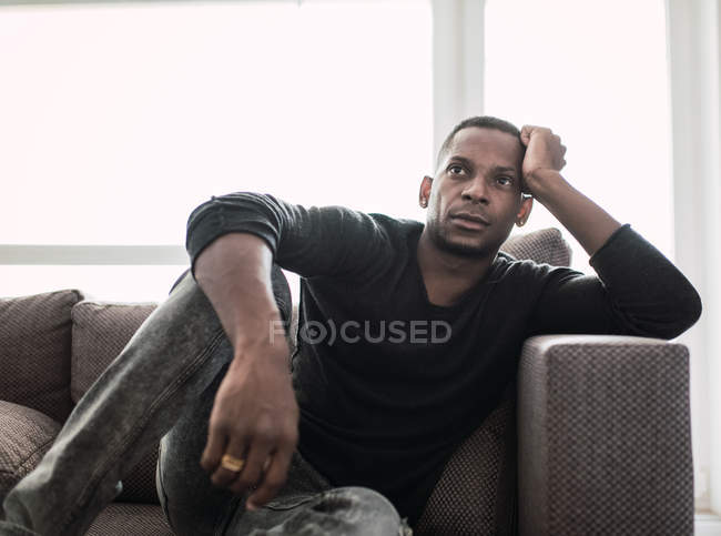 Мечтающий взрослый черный мужчина, опирающийся на руки, сидя на диване и глядя в блестящее окно — стоковое фото