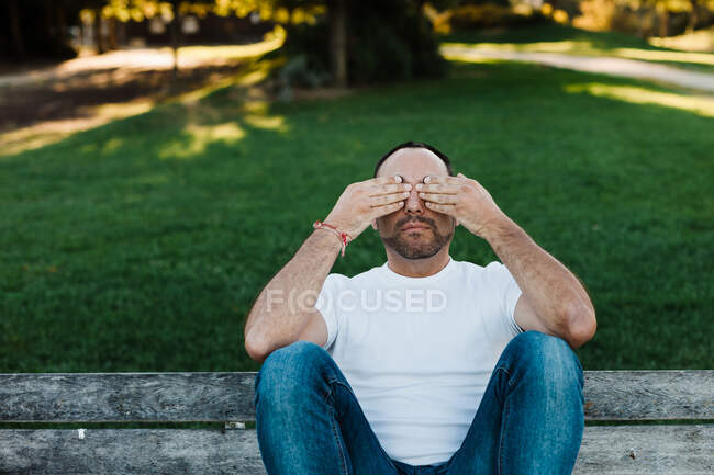 Attractive adult male covering eyes while sitting on bench in park on sunny day — Stock Photo