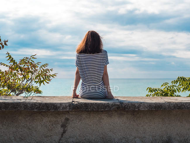 Странствующая женщина сидит на каменном заборе на фоне облачного неба и синего моря — стоковое фото