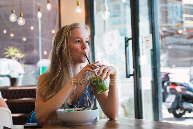 Bella donna bionda che beve frullato verde mentre si rilassa da solo in caffè con ciotola di cibo vicino — Foto stock