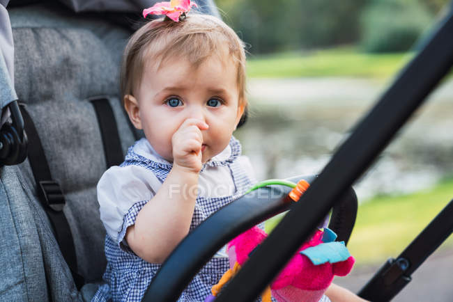 Adorable baby girl looking at camera and sucking thumb while sitting in carriage on blurred background of park — Stock Photo