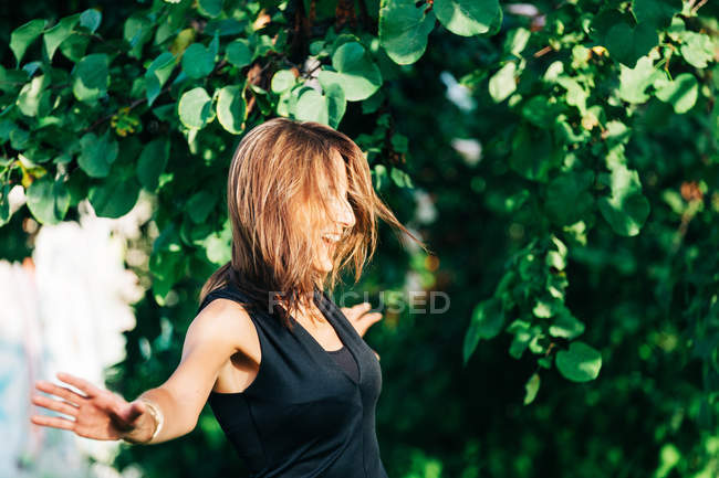Cheerful stylish woman standing under tree and smiling at camera in park — Stock Photo