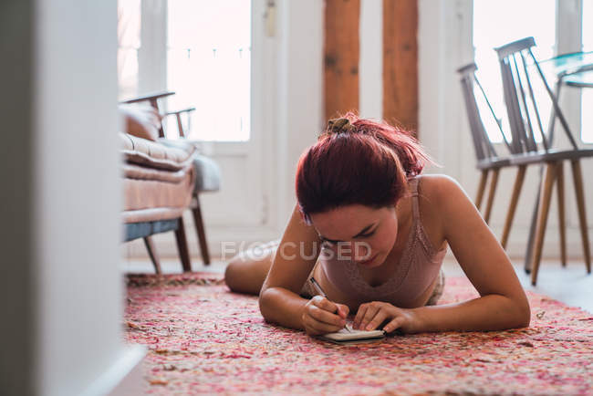 Женщина в нижнем белье лежит на полу и пишет в блокноте — стоковое фото