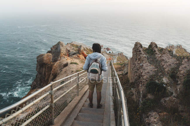 Rückansicht eines Mannes mit Rucksack, der auf Metallstufen auf einer steinigen Klippe steht und einen bewundernswerten Blick auf das herrliche Meer in Point Riyes, Kalifornien hat — Stockfoto