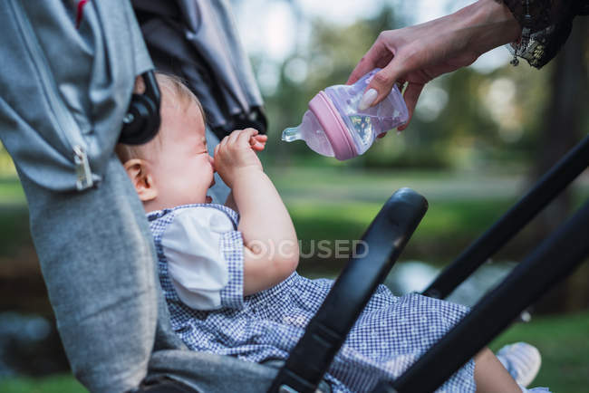 Mano de madre sosteniendo botella de agua frente a bebé llorando en cochecito sobre fondo borroso del parque - foto de stock
