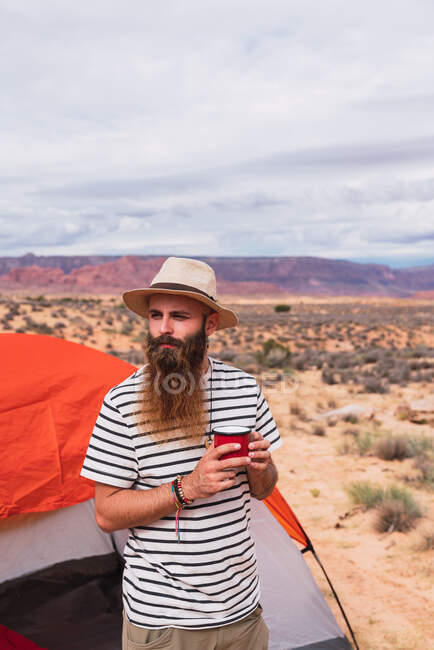 Бородатий хлопець у повсякденному вбранні тримає кухоль гарячого напою та сучасний смартфон, сидячи біля намету та дивлячись у чудову природу — стокове фото