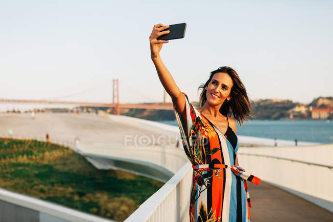Femme en robe d'été colorée en utilisant le téléphone et en prenant selfie du pont — Photo de stock