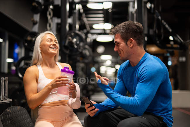 Hombre y mujer en forma alegre sentados en el gimnasio y riendo - foto de stock