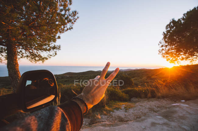 Mano di uomo anonimo che mostra V-segno vicino specchio ala contro maestosa campagna durante bel tramonto — Foto stock