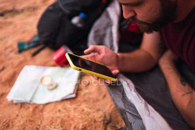 Урожай бородатий чоловік їсть свіже яблуко і переглядає сучасний смартфон, лежачи в наметі під час кемпінгу в пустелі — стокове фото