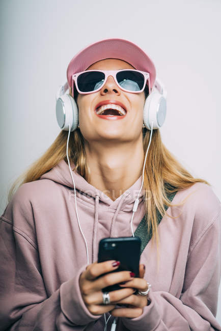 Stylish woman in pink hoodie and cap listening to music with headphones and laughing — Stock Photo