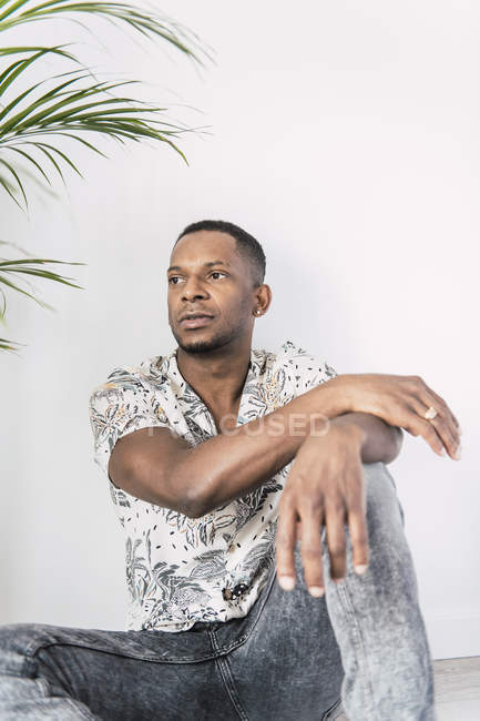 Homme noir pensif assis sur le sol et regardant loin contre le mur blanc avec la plante verte — Photo de stock