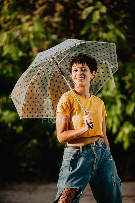 Молода жінка в стильному повсякденному вбранні дивиться на камеру, тримаючи парасольку і стоячи на вулиці біля кущів — стокове фото