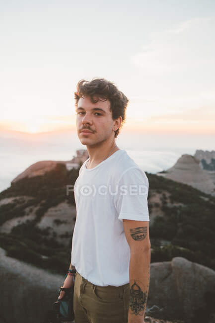 Vue latérale d'un gars attrayant avec une moustache tenant un appareil photo et regardant un appareil photo debout sur une falaise lors d'un beau lever de soleil à Barcelone, Espagne — Photo de stock