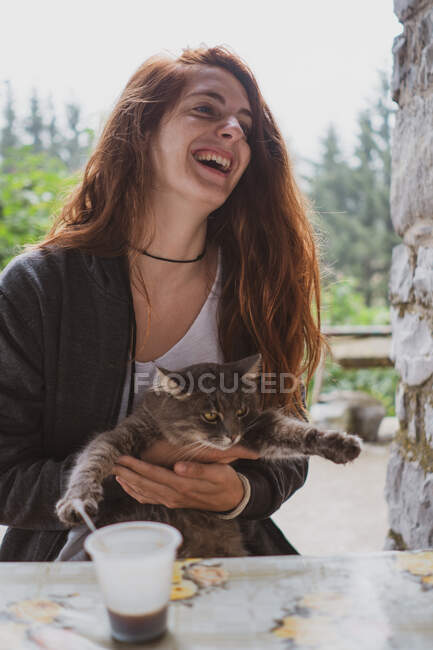 Schöne junge Frau hält süße Katze und lacht, während sie am Tisch in der Nähe von ländlichen Gebäude in Bulgarien, Balkan sitzt — Stockfoto