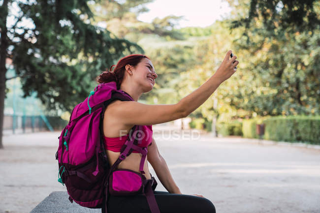 Mujer joven sonriente en ropa deportiva con mochila rosa sentada en el parque y tomando selfie con teléfono inteligente - foto de stock