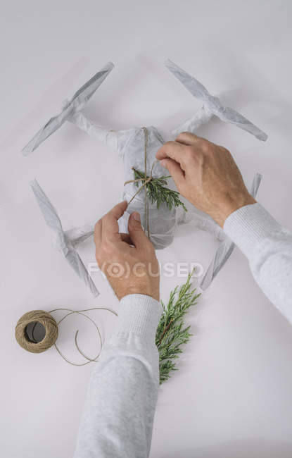 Manos masculinas decorando dron envuelto como regalo de Navidad con rama de abeto y cordel sobre fondo blanco - foto de stock