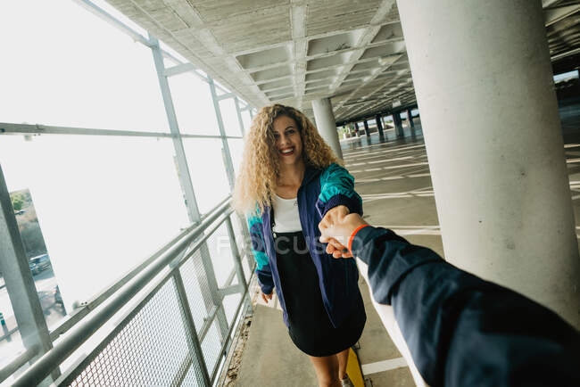 Прекрасна молода жінка з кучерявим волоссям посміхається і тримає руку анонімного друга, пропонуючи слідувати за нею на даху — стокове фото