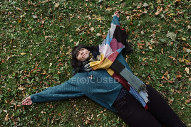 From above of brunette in sweater and colorful scarf lying on meadow with hands apart in autumn looking dreamily up — Stock Photo