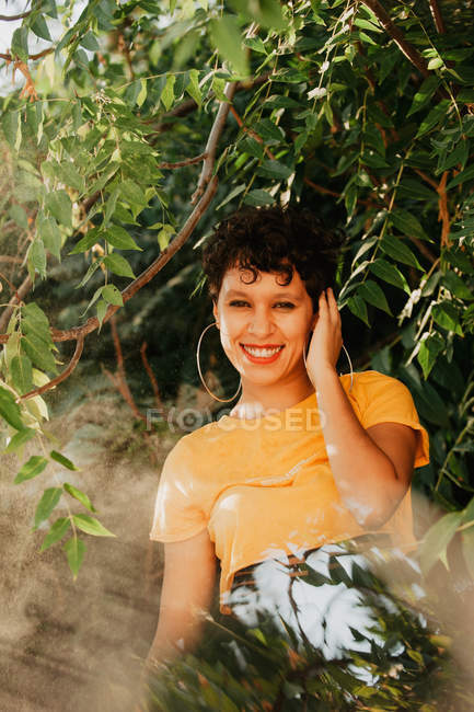 Retrato de morena sonriente con el pelo corto de pie en la vegetación verde con luz solar - foto de stock