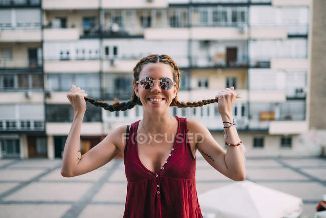 Весела руда молода жінка в сонцезахисних окулярах тримає коси проти житлового будинку — стокове фото
