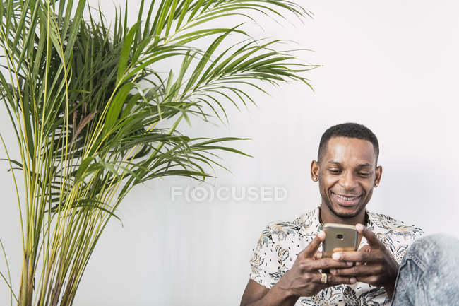 Homme noir souriant utilisant un smartphone contre un mur blanc avec une plante verte — Photo de stock