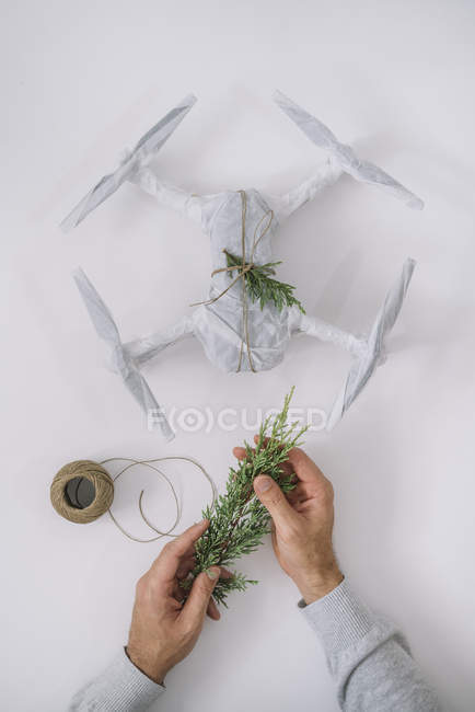 Mains masculines décorant drone enveloppé comme cadeau de Noël avec branche de sapin et ficelle sur fond blanc — Photo de stock