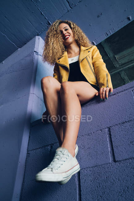 Elegante donna bionda riccia in scarpe da ginnastica e giacca gialla seduta sul muro viola e ridendo — Foto stock
